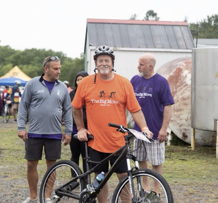 A rider in a Big Move Cancer Ride shirt with his bicycle, with supporters alongside